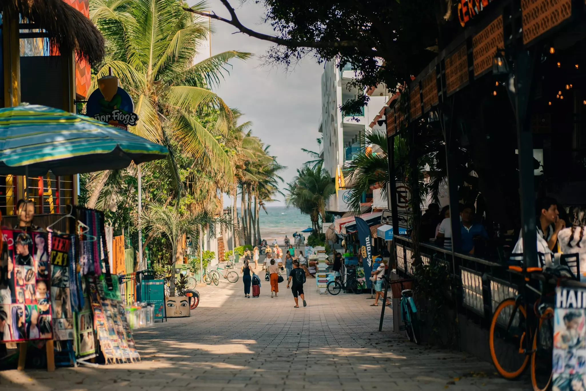 People walk down a pedestrianized street toward the beach in a seaside town. Palm trees and businesses line the street.