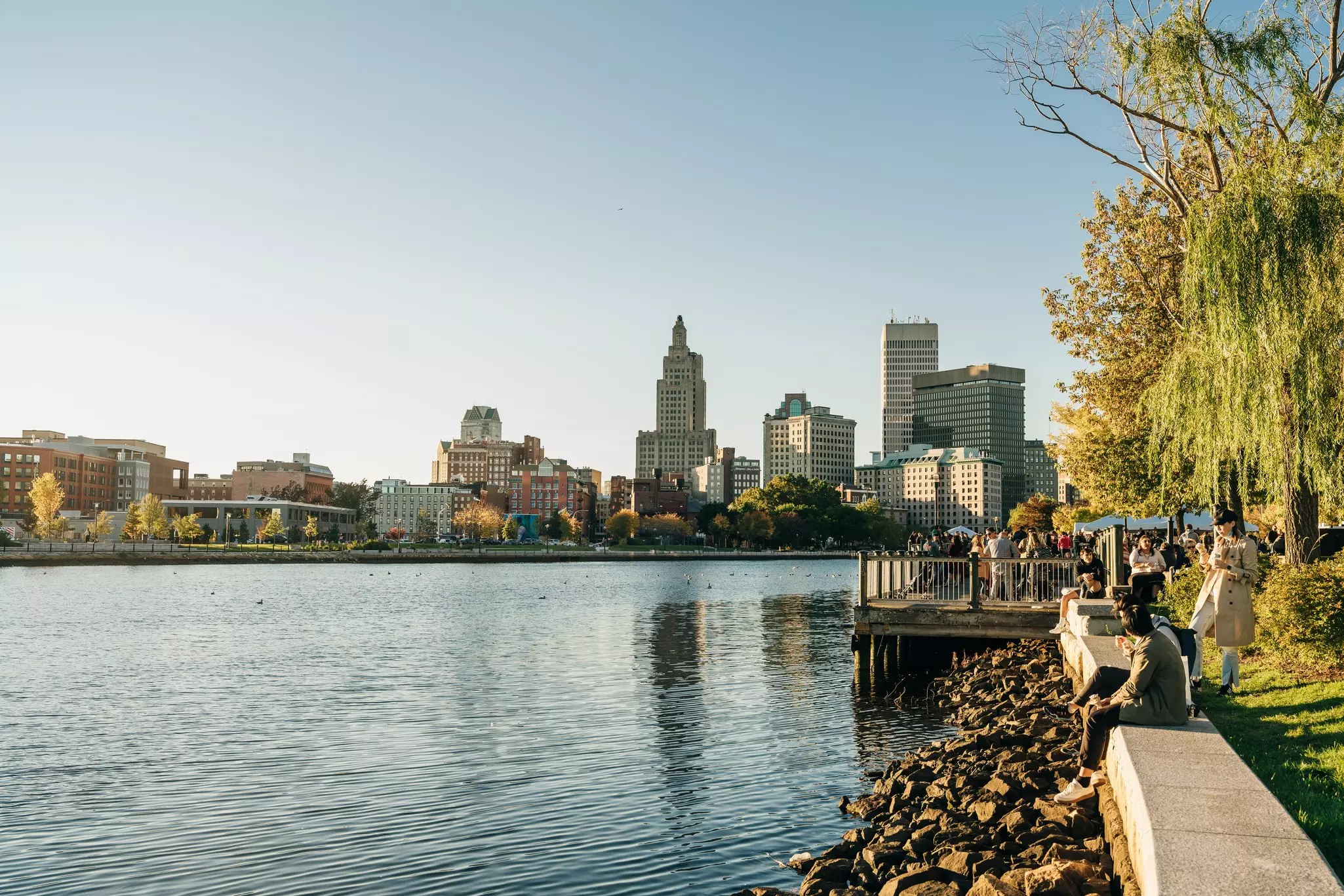 View of the Providence, Rhode Island, skyline from across a river; people are sitting on a wall by the opposite shore and standing on a deck over the water.