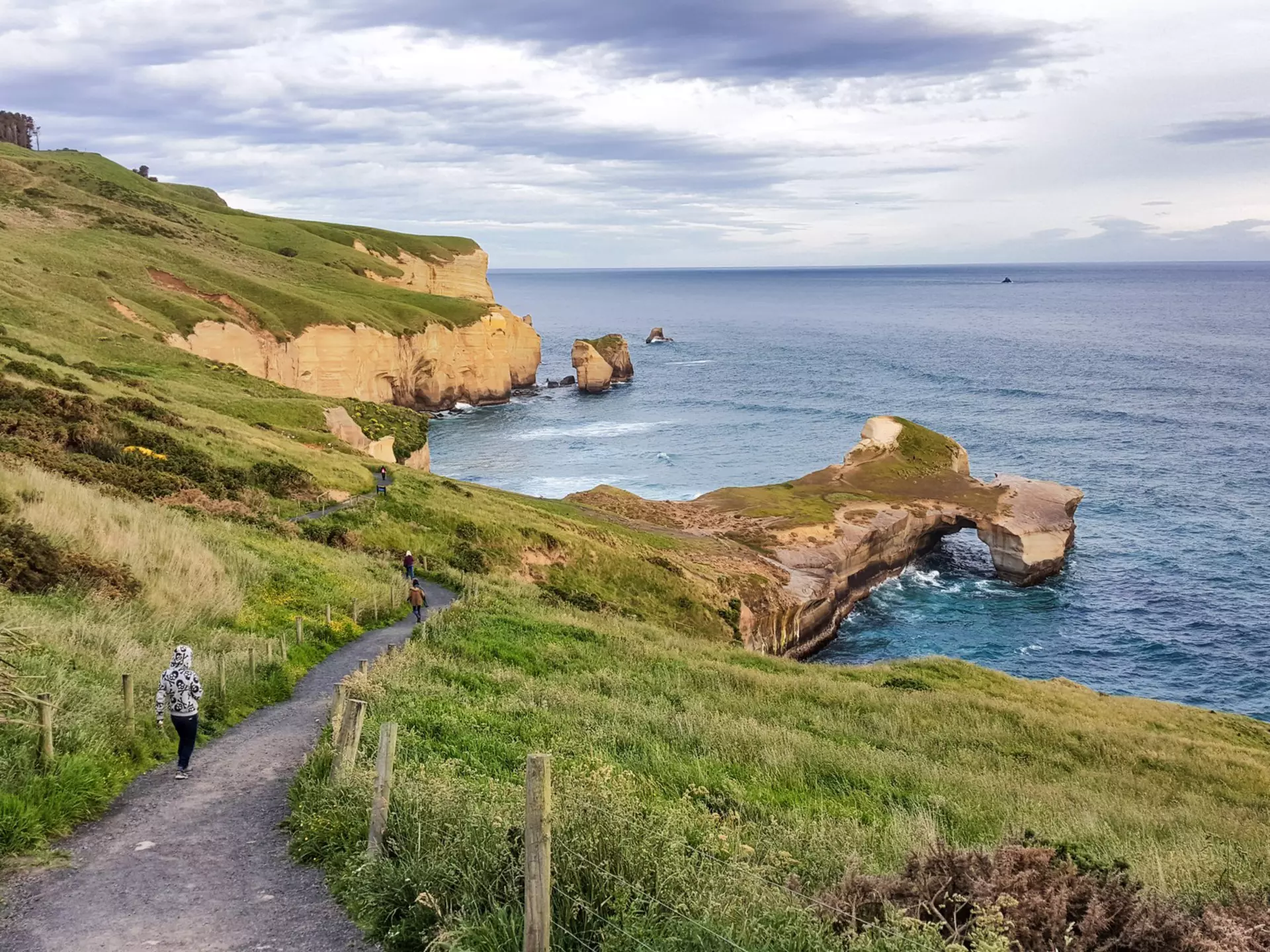 People wander down a steep cliff path towards a beach, with islets and a rock arch.