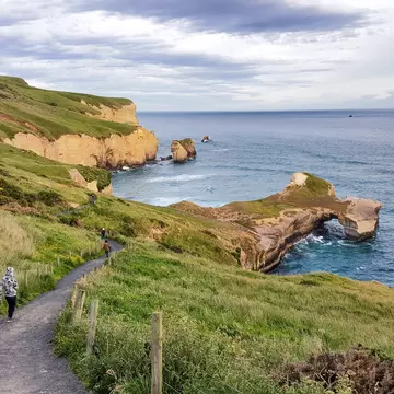 People wander down a steep cliff path towards a beach, with islets and a rock arch.
