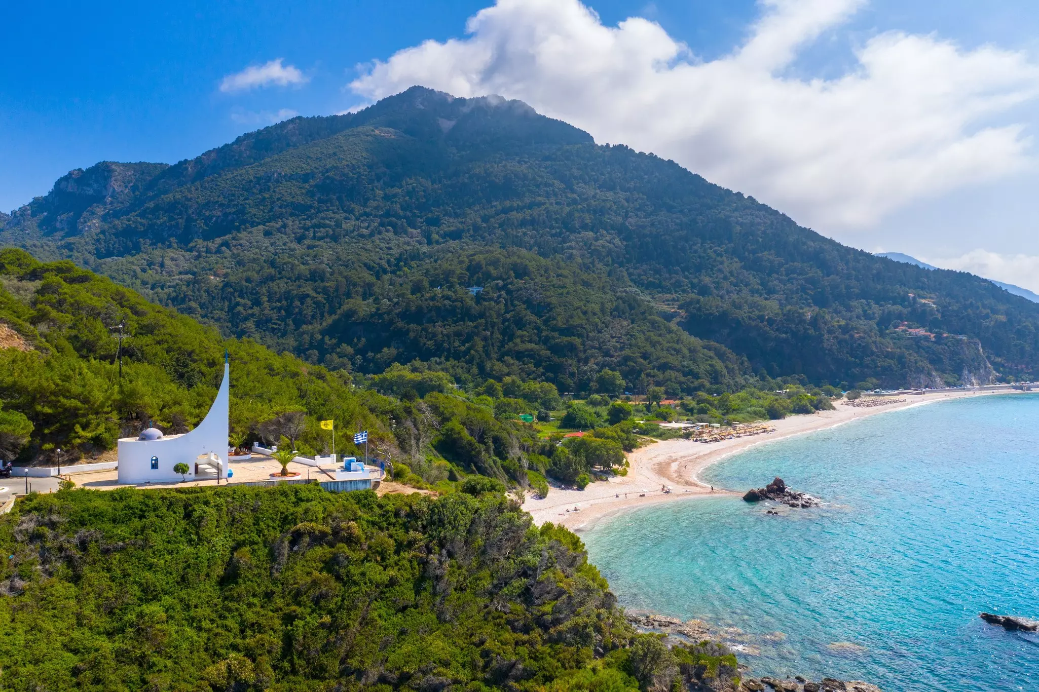 A white modern building is seen atop a tree-covered hill. A beach and the sea are visible in the distance.