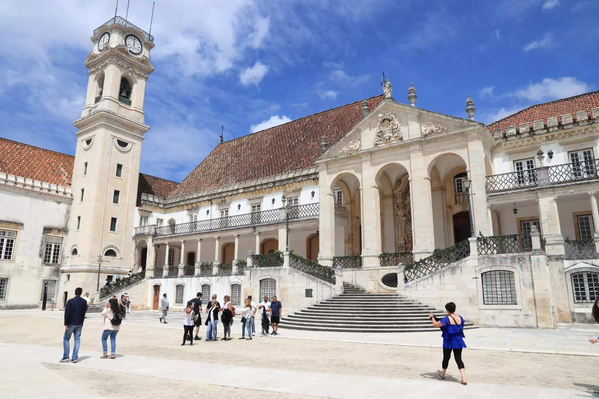 People touring white flagstone courtyard near stone buildings with reddish roofs on a sunny day.