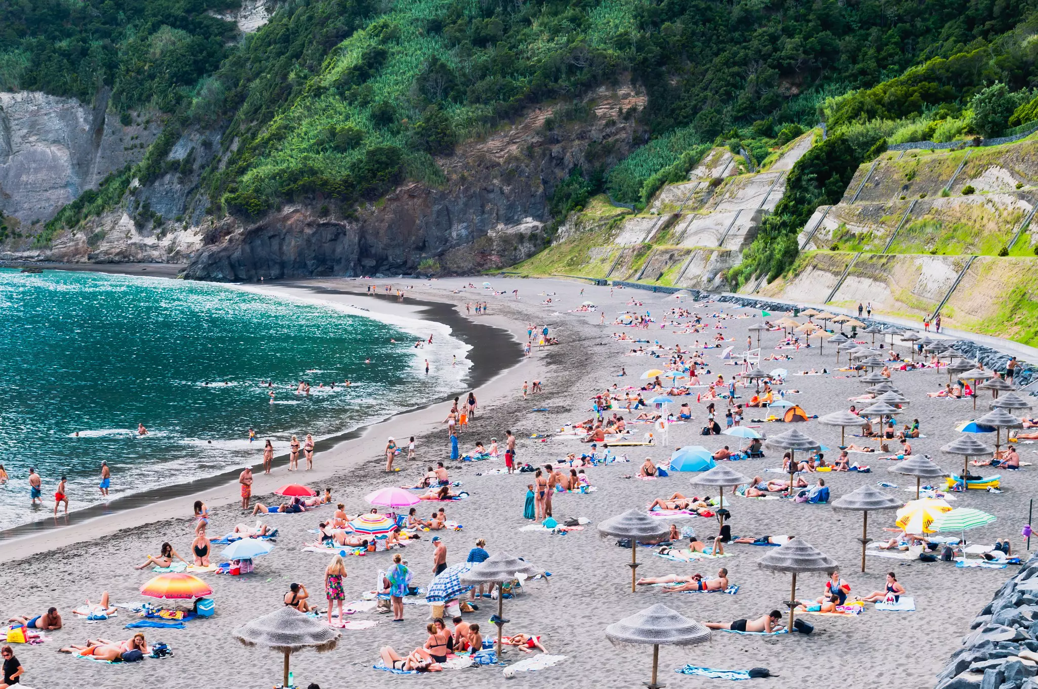 Sunbathers and colorful umbrellas on a gray sand beach backed by green cliffs.