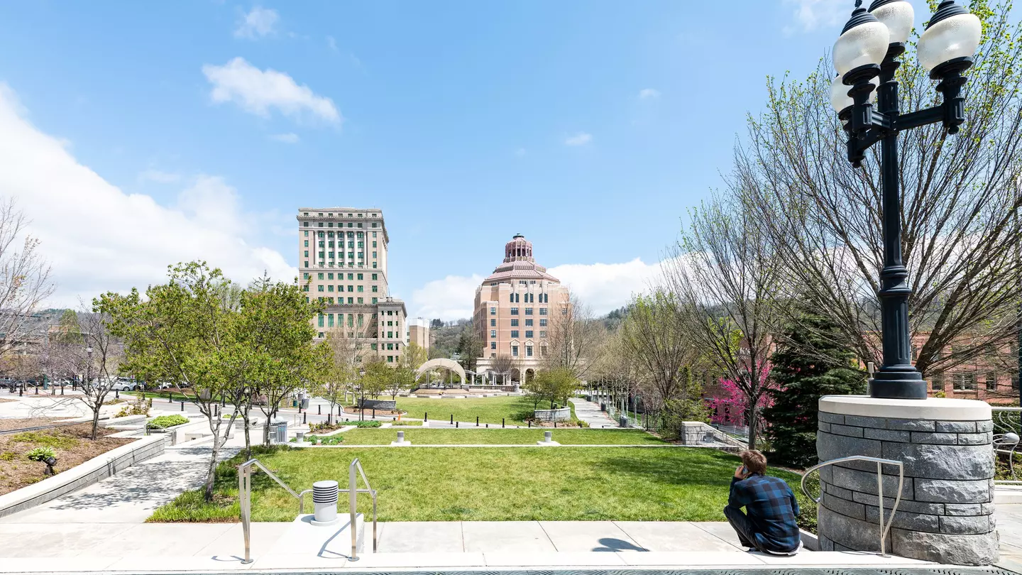 Back of young hipster millennial man sitting looking at buildings, courthouse and green Pack Square Park in hippie North Carolina NC famous town, city in the mountains, talking on phone
