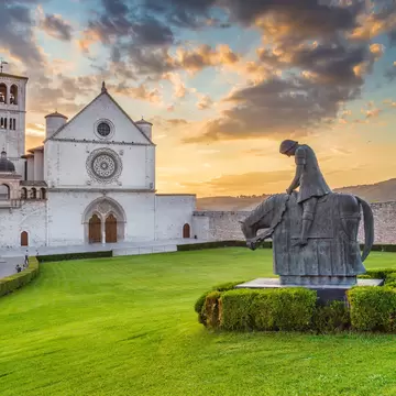 White stone facade and bell tower of the Basilica of St Francis in Assisi at sunset, with a bronze knight statue on the lawn