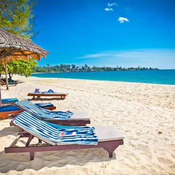 Wooden sun loungers with striped towels under a palm-thatch parasol on white sand, turquoise water and a wooded headland beyond.