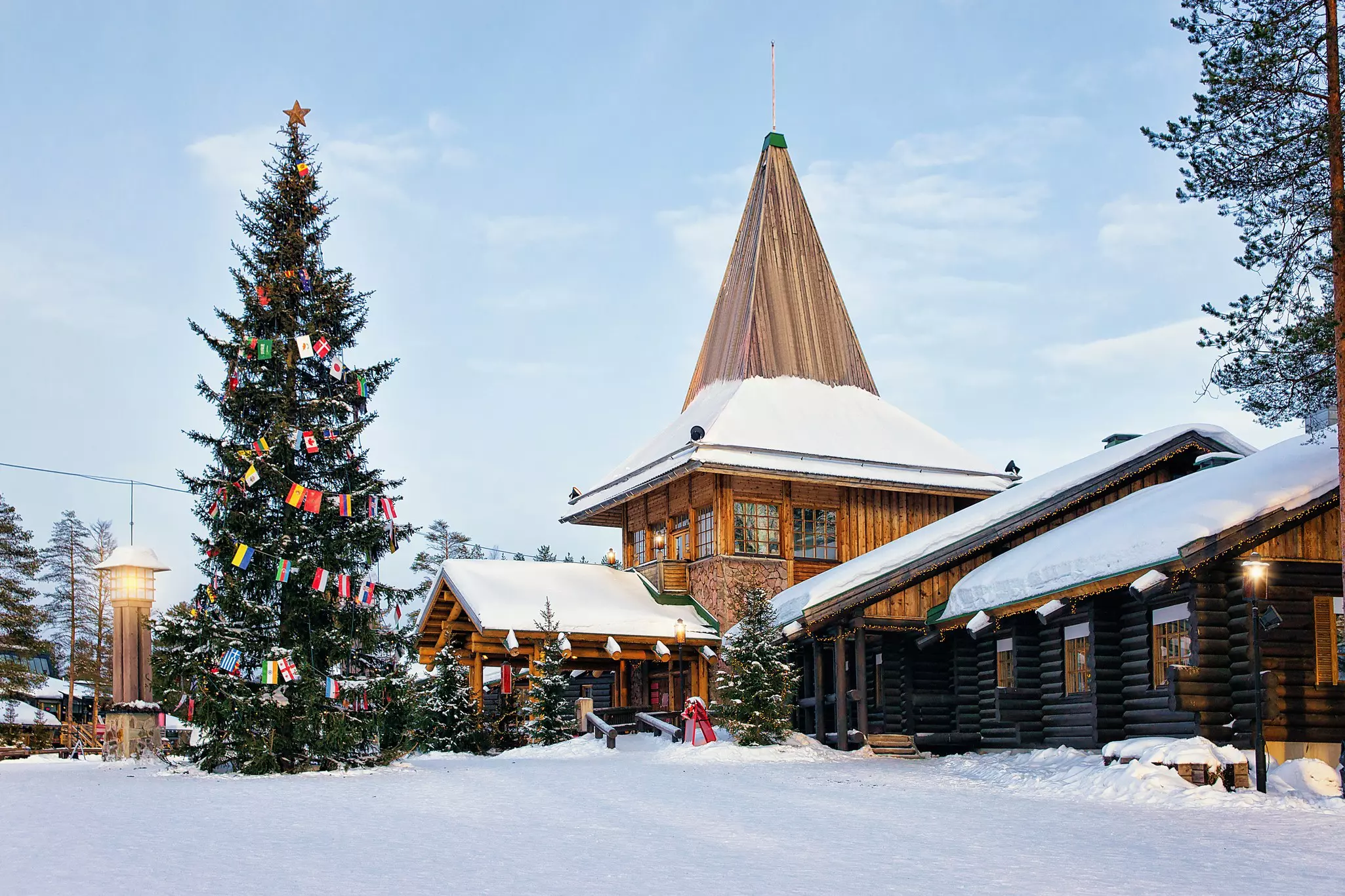 A stone and wood building with a steep peak; there is snow on the roof and ground and an evergreen tree decorated with flags.