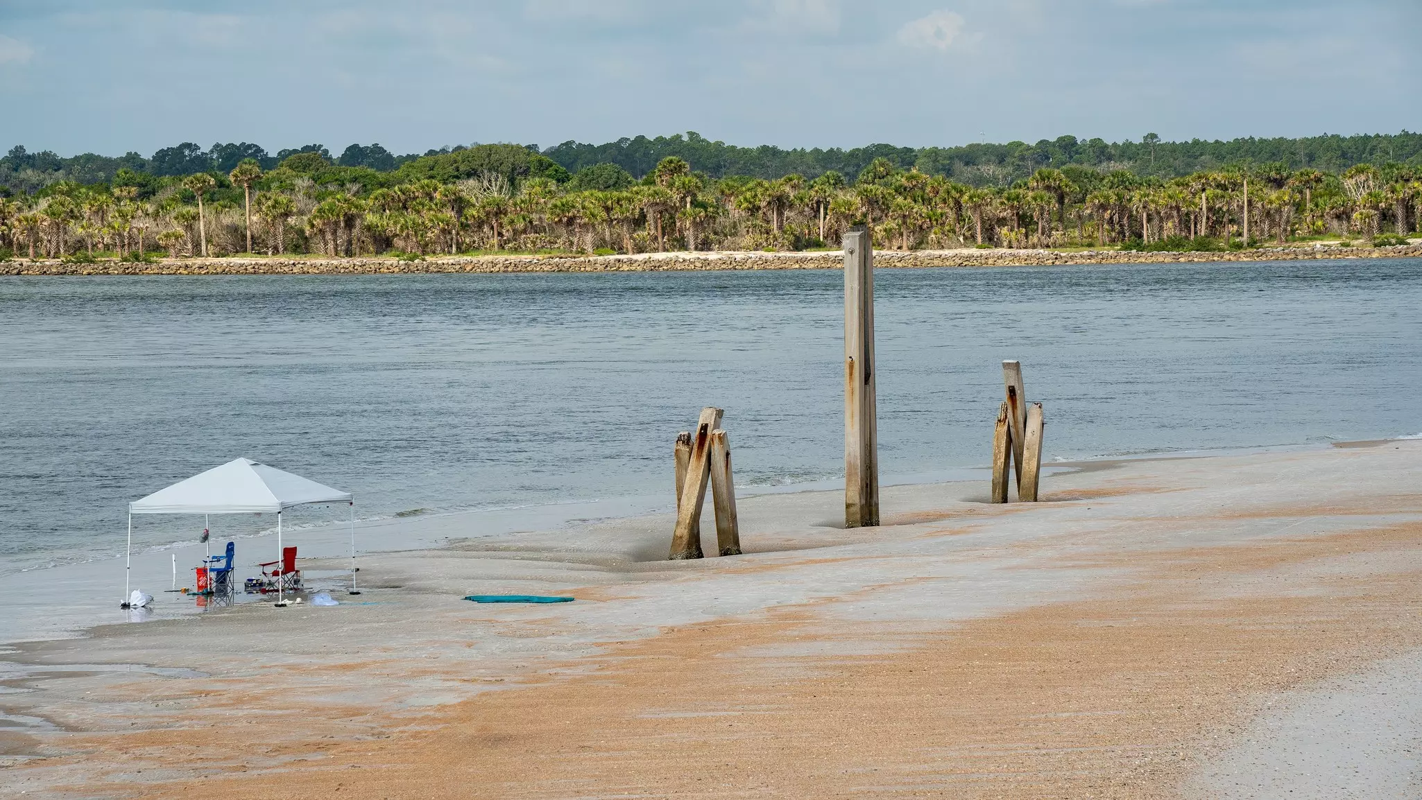 Two people under a tent sit by the on the beach of an inlet, fishing.