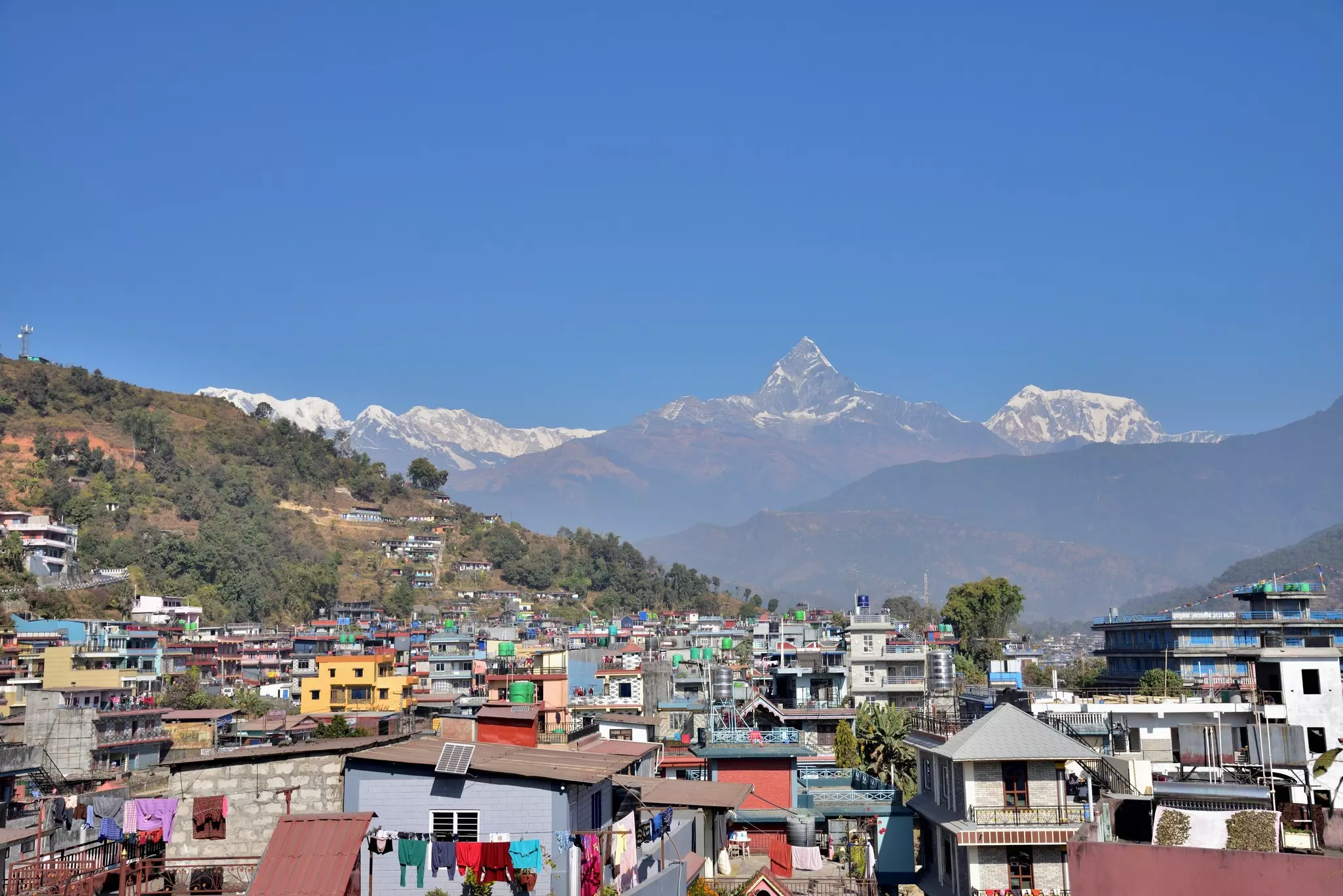 A cluster of buildings in Nepal, with snowcapped peaks in the background under blue sky.