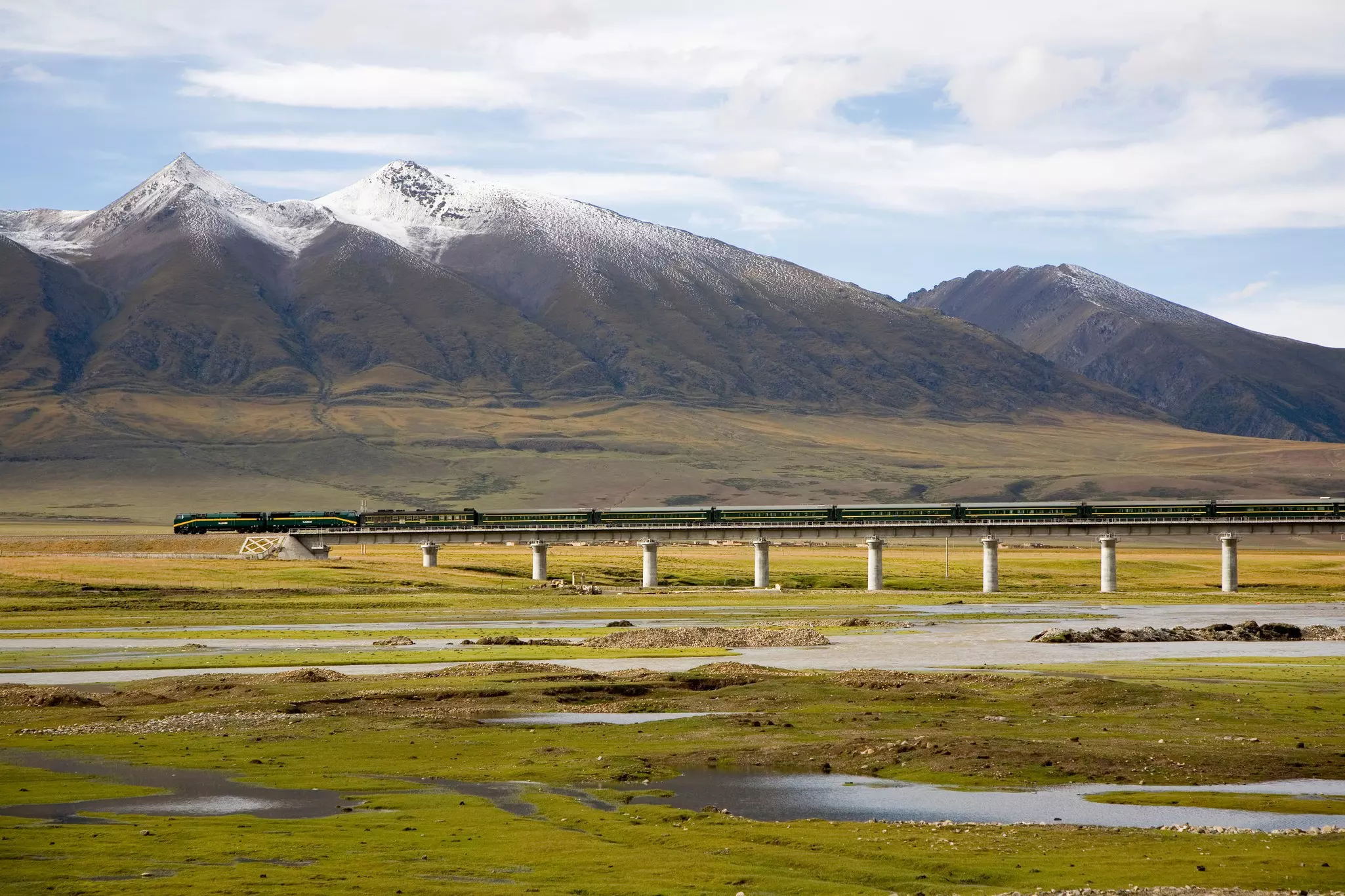 A train crosses a low bridge over a wetland backed by snowcapped mountains.