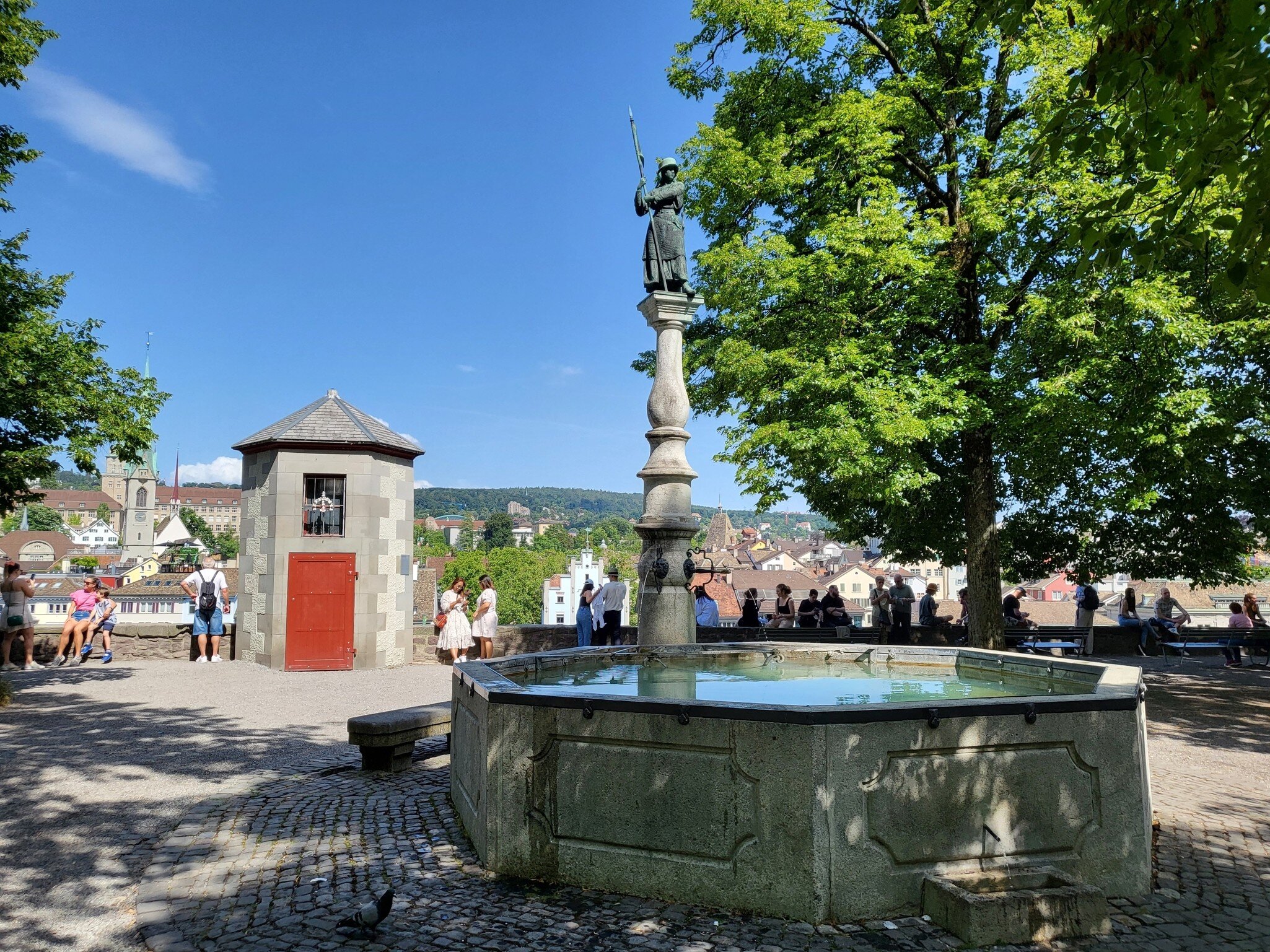 Stone fountain topped by a bronze figure with a spear, a big leafy tree and visitors at a low wall overlooking rooftops