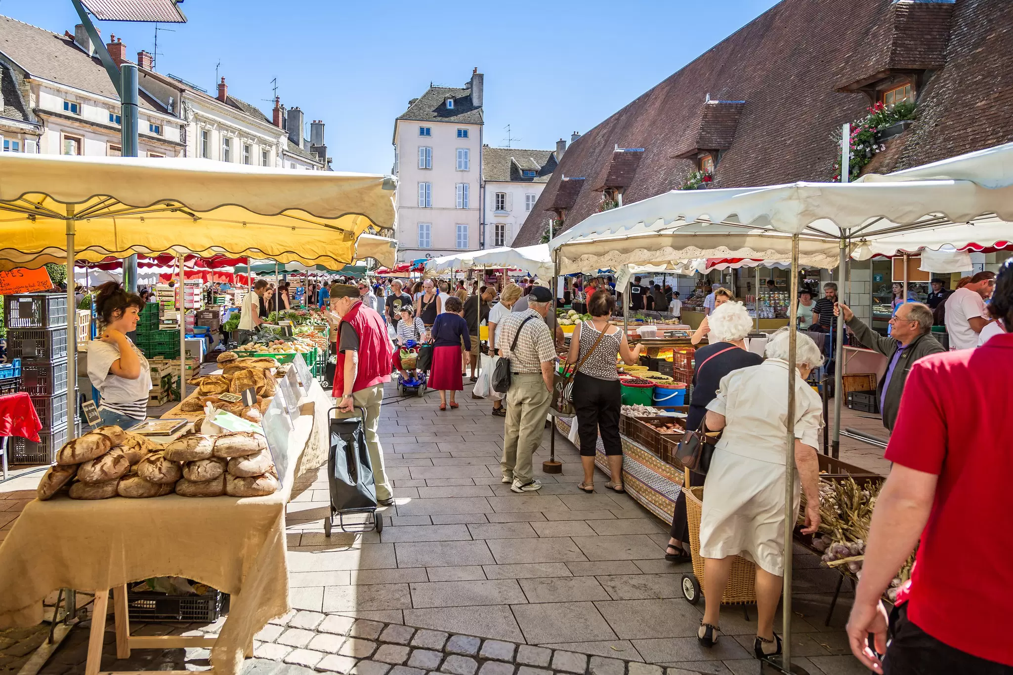 People shop for vegetables and other produce at market stalls set up in a town square.
