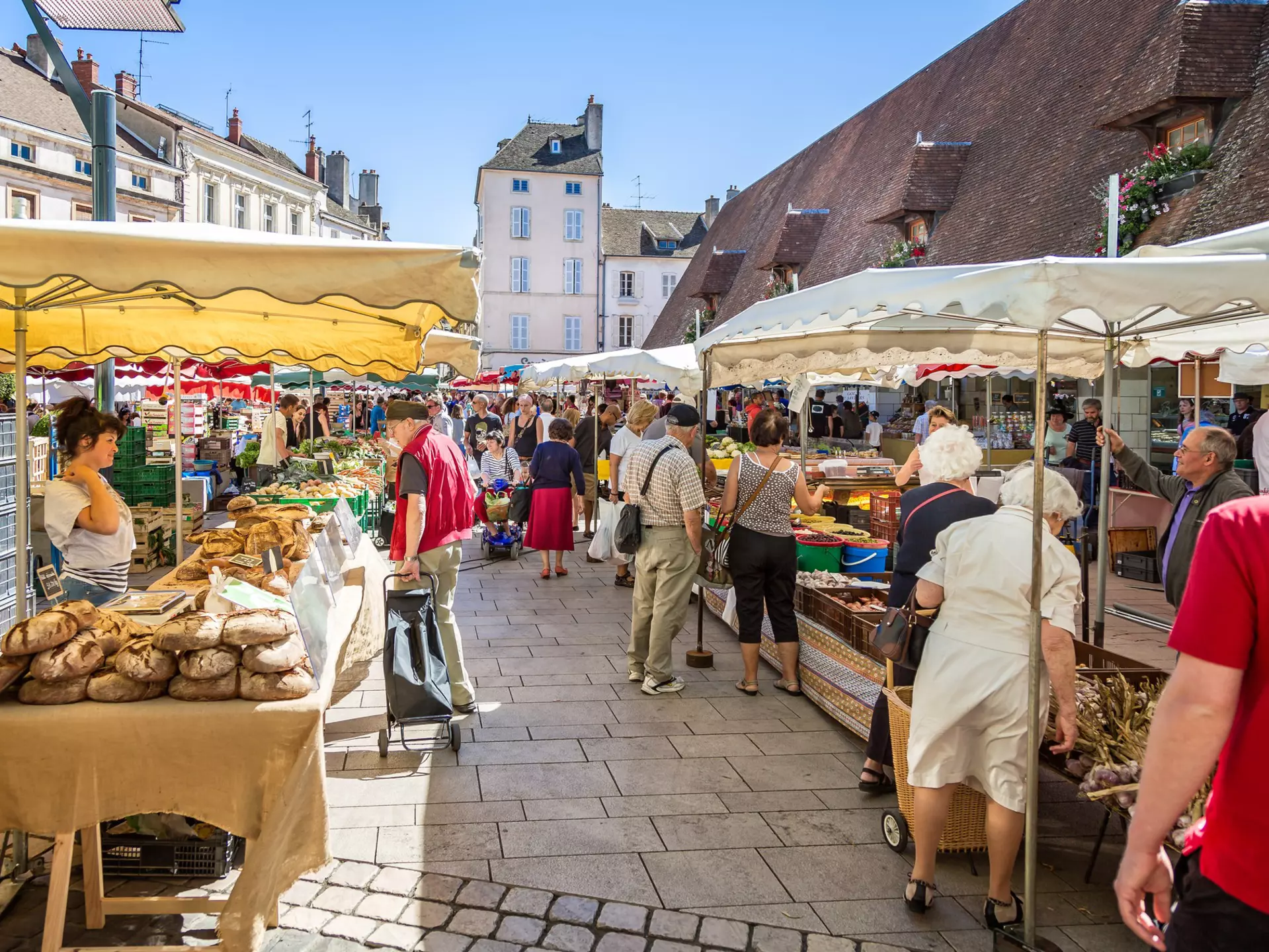 People shop for vegetables and other produce at market stalls set up in a town square.