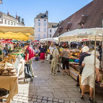 People shop for vegetables and other produce at market stalls set up in a town square.