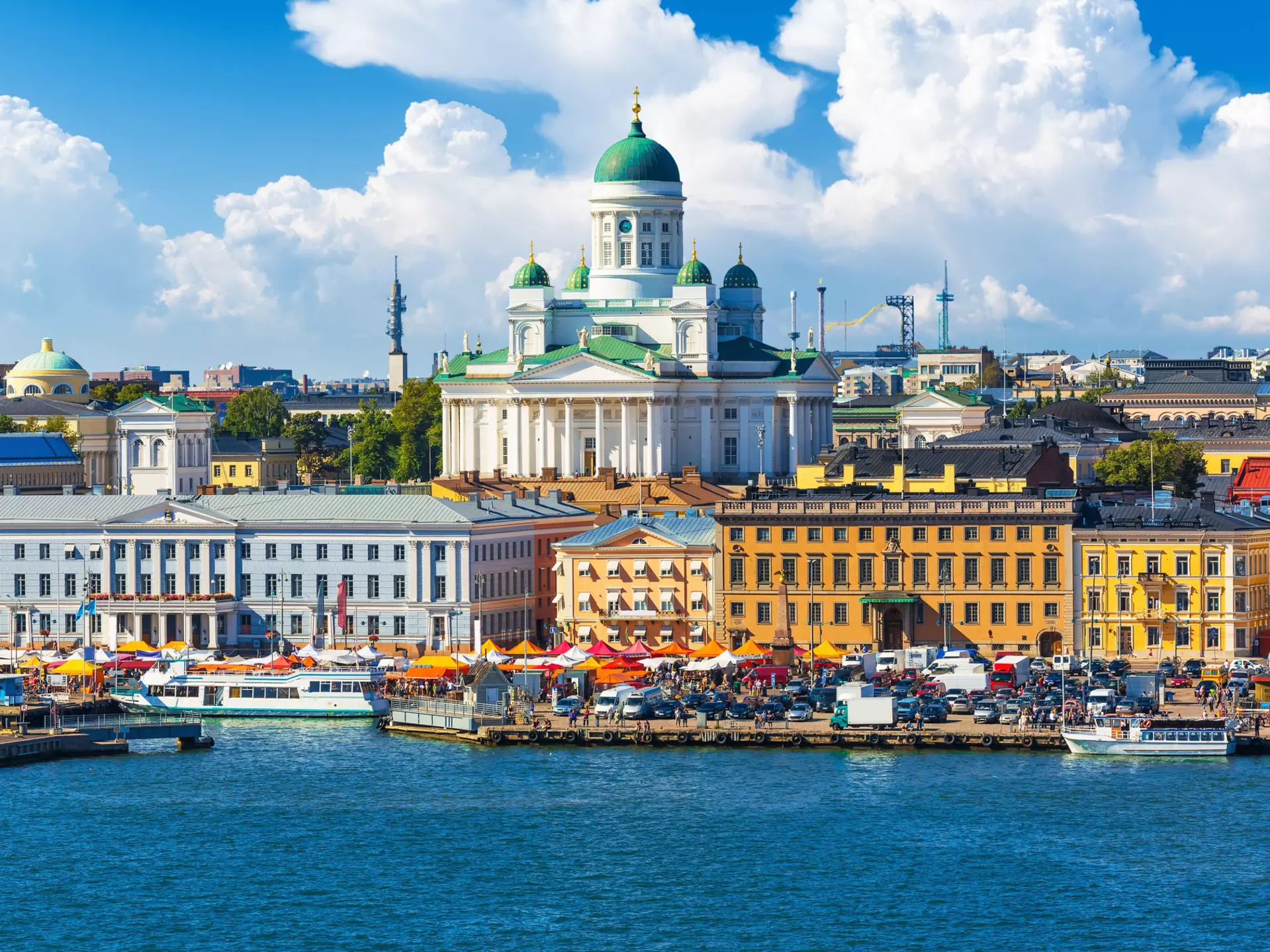 A city waterfront in summer, with people sat under sunshades in Helsinki, Finland. A large cathedral looms above.