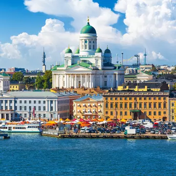 A city waterfront in summer, with people sat under sunshades in Helsinki, Finland. A large cathedral looms above.