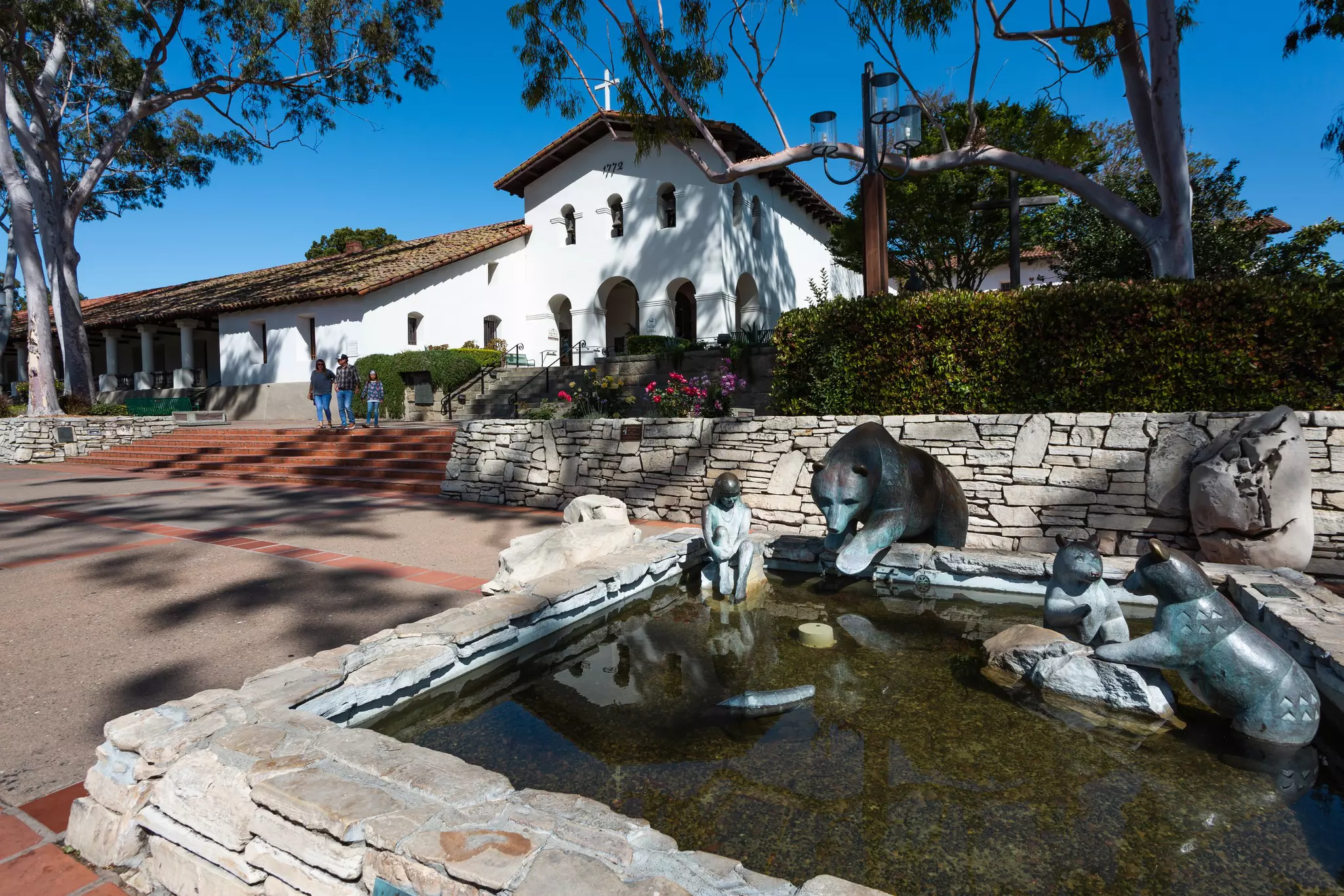 A sculpture in a fountain depicts children and bears in an outdoor plaza. A white mission building is visible in the background.