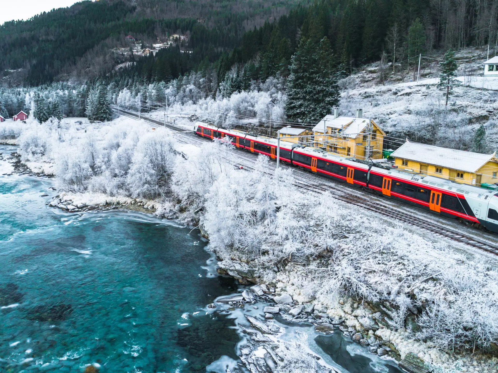 A red train travels through snowy surrounds, with yellow buildings on the right and a frigid lake to the left of frame.