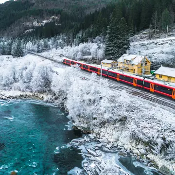 A red train travels through snowy surrounds, with yellow buildings on the right and a frigid lake to the left of frame.