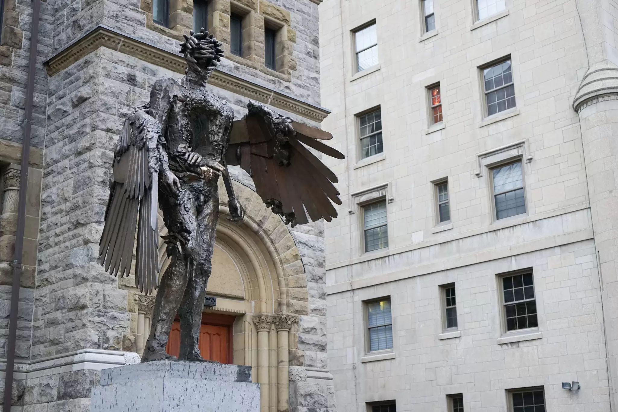 A large metallic sculpture of a human form with wings stands on a plinth outside a building