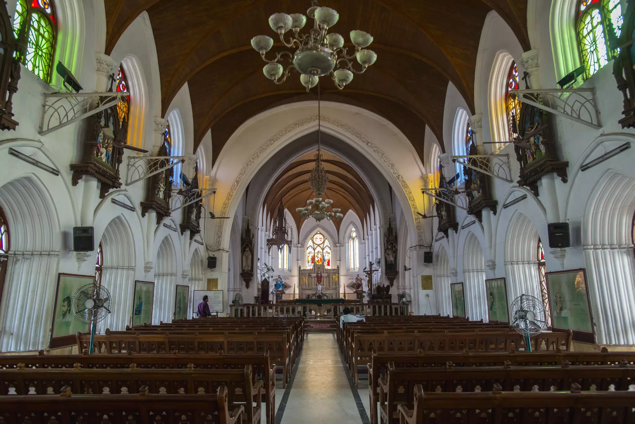 Interior of a cathedral, looking directly down the central aisle. There are dark wooden pews on either side and grand white arches overhead.