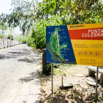 A large painted welcome sign with a site map beside a paved path through dry tropical scrub, a stone bench alongside.