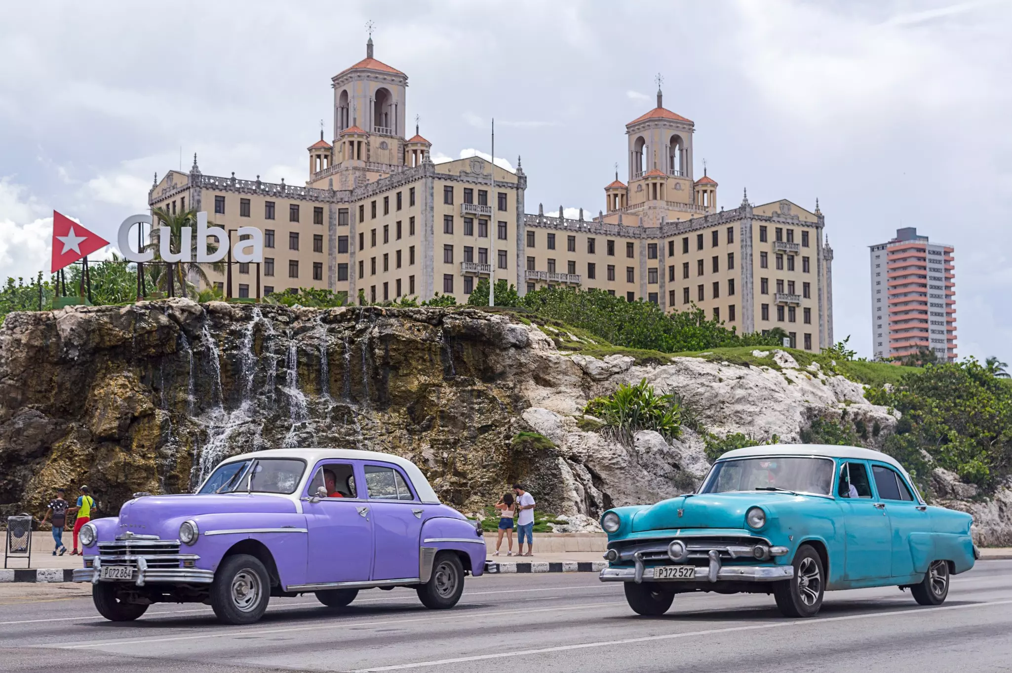 Two vintage cars drive along a city road. A grand hotel is seen over cliffs next to the road.