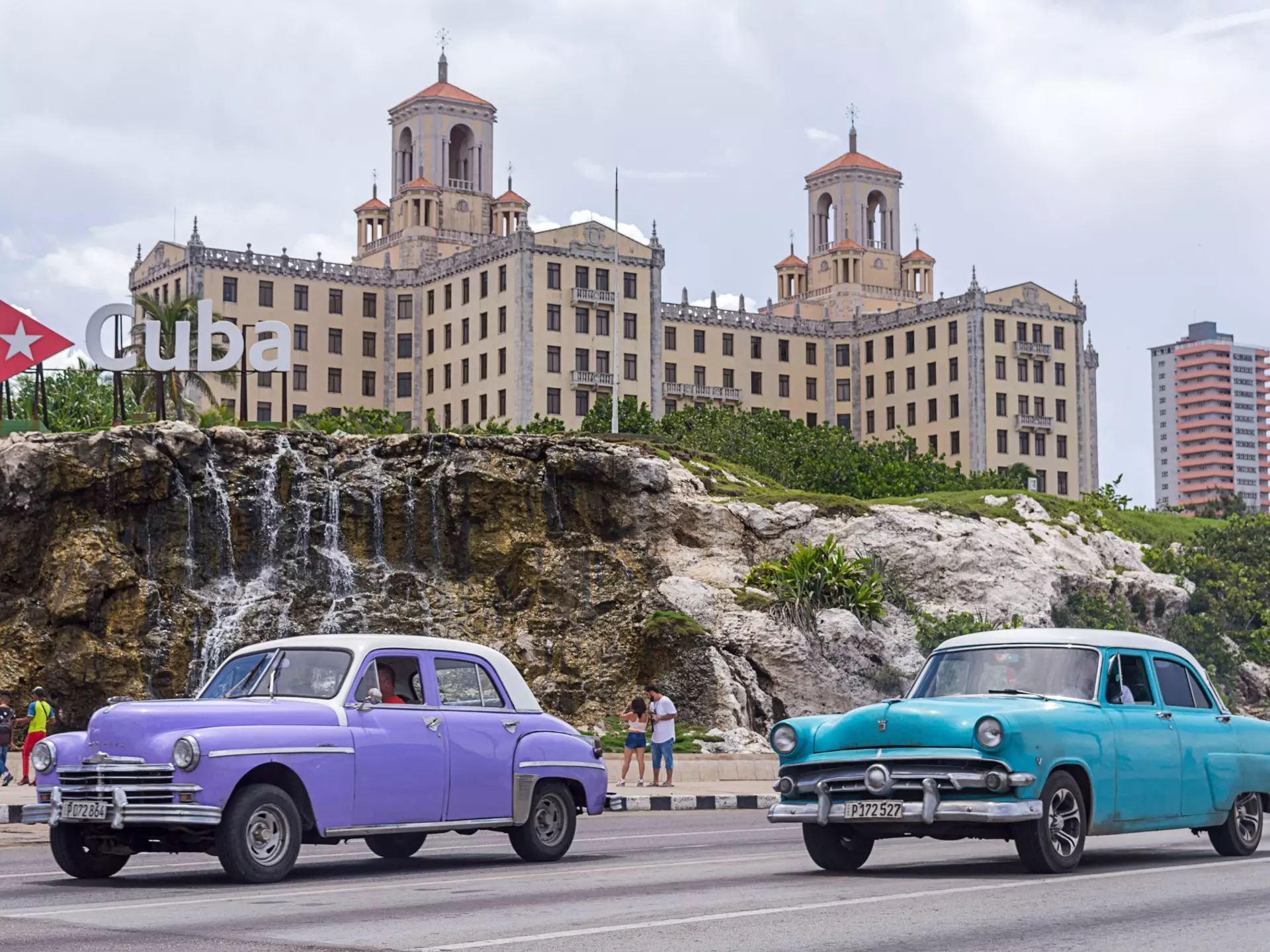 Two vintage cars drive along a city road. A grand hotel is seen over cliffs next to the road.