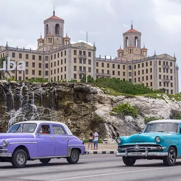 Two vintage cars drive along a city road. A grand hotel is seen over cliffs next to the road.