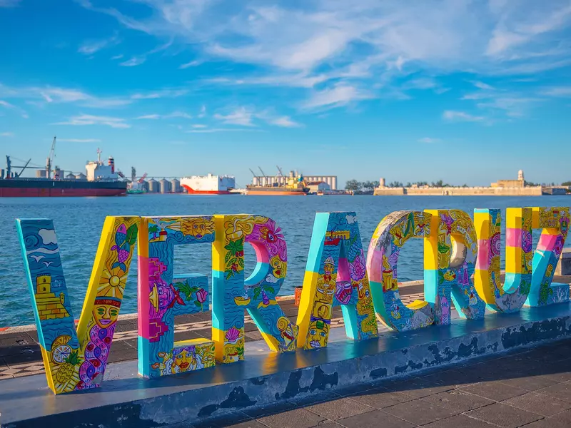 A colorfully painted sign of tall letters spelling "Veracruz" on a waterfront promenade overlooking a port wih cargo ships in the background on a sunny day.