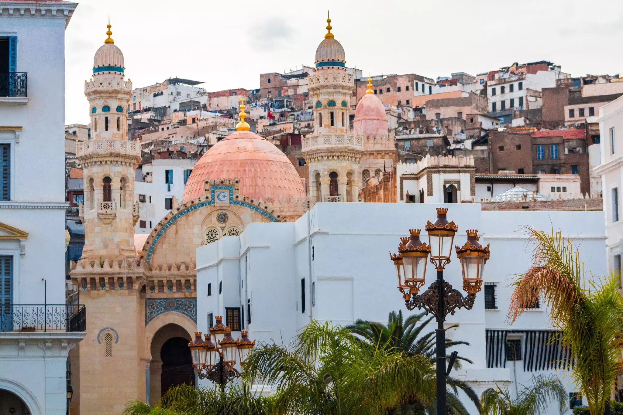 The towers of a mosque in Algiers appear in the foreground, with a hillside of historic buildings rising behind it.
