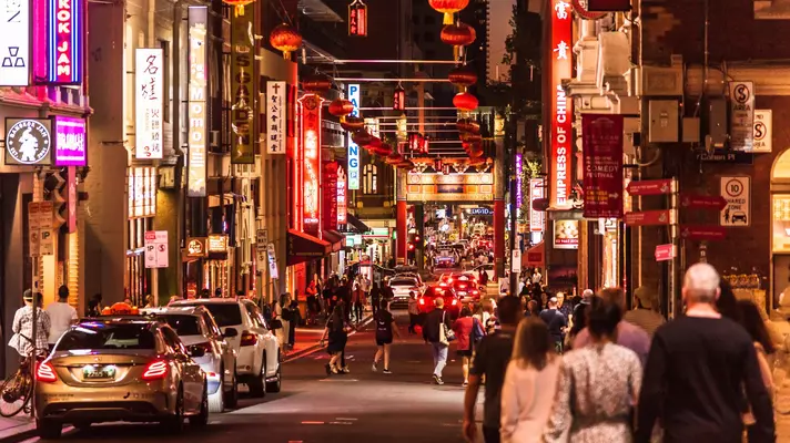 People and cars on a street in Melbourne with illuminated signs at night.
