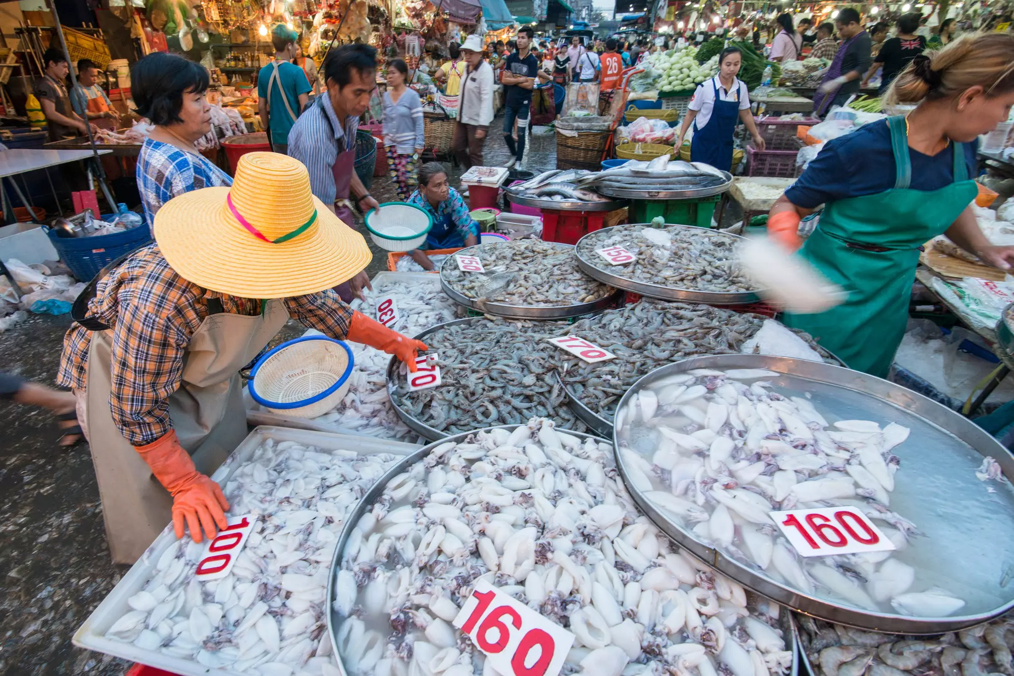 Women wearing gloves and oversized hats sort through large plates of fish at a fish market