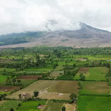 Cloud-capped volcano with grey ash-covered slopes behind a patchwork of green fields and scattered houses
