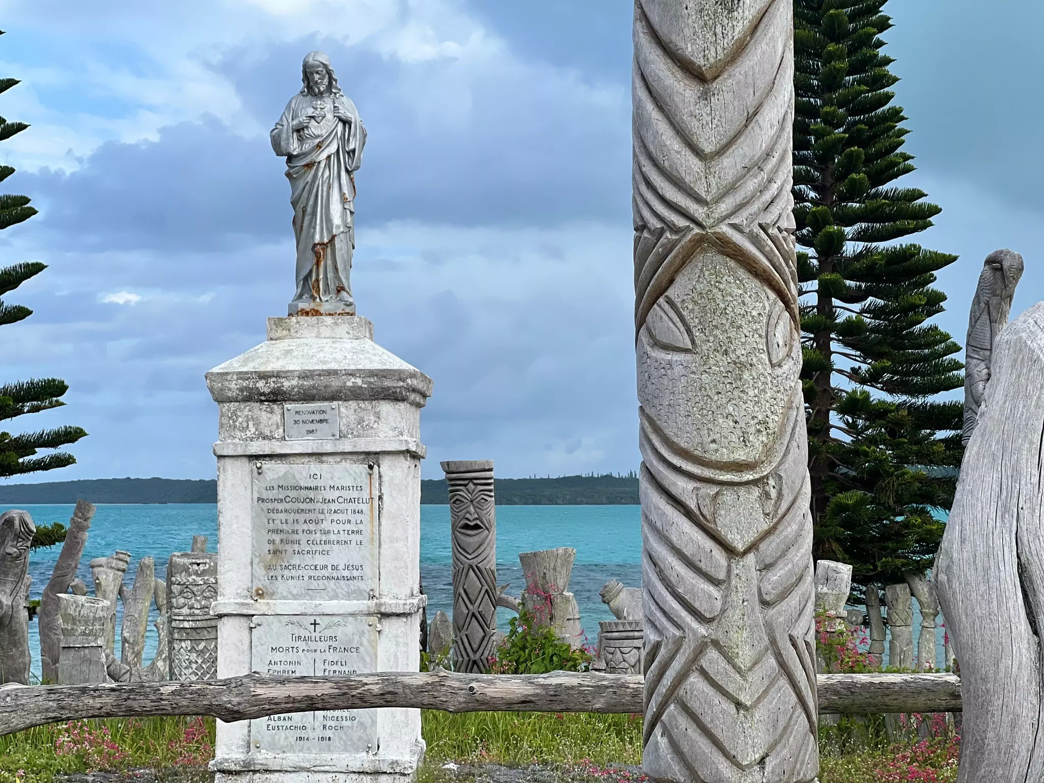 A series of carved totem poles stand near a monument topped with a statue of Christ beside the ocean.