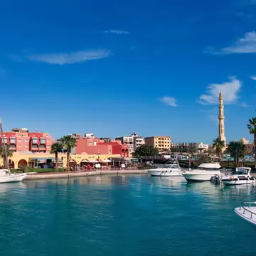 A small harbor with several docked yachts and small motorboats. A mosque with two tall minarets stands on the shore.