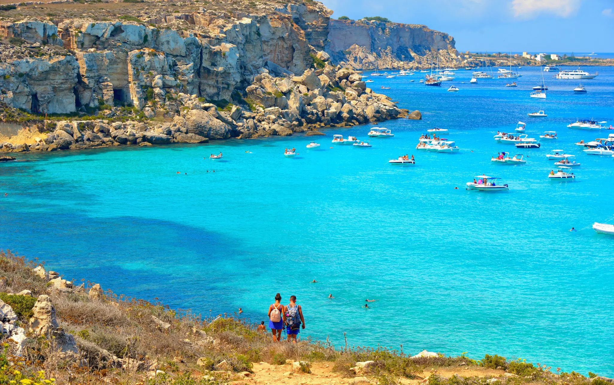 Two people are seen from behind walking on a hill toward a cove with bright blue water where boats are moored.