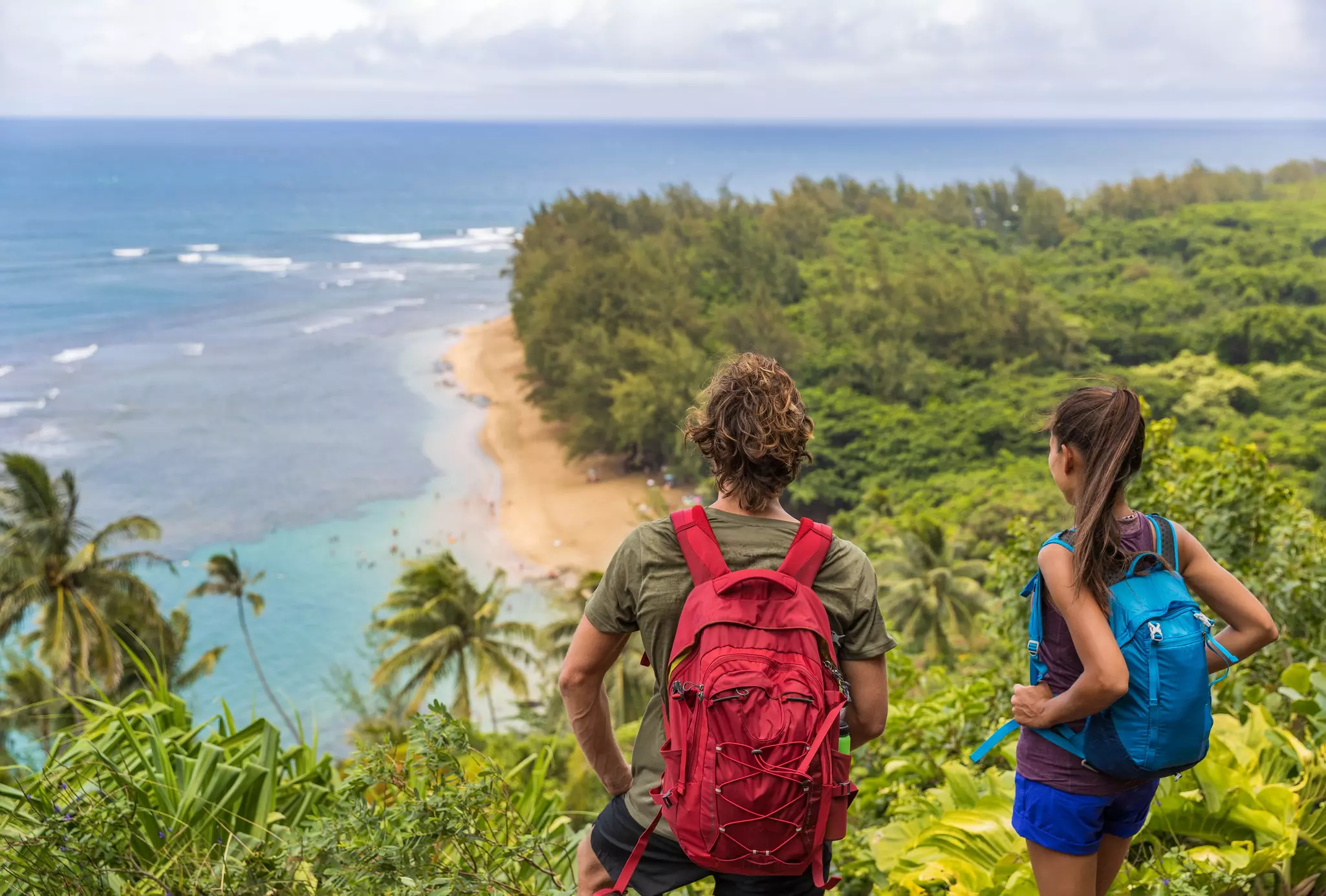 Hikers couple hiking on Kalalau trail overlooking beach coastal walk two tourists with backpacks walking outdoor in Kauai island, Hawaii.