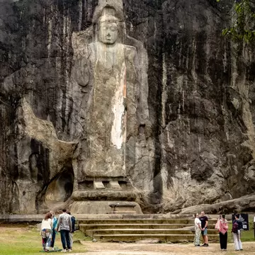 Tourists standing in front of the Buduruwagala Raja Maha Viharaya in Sri Lanka, an ancient Buddhist temple with massive rock carvings of Buddha, surrounded by forest.