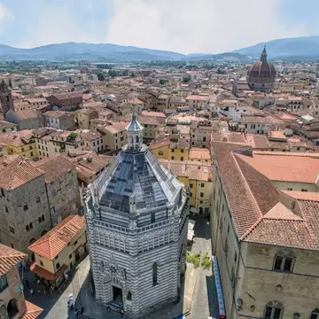 An aerial view over Pistoia, Italy, centred on an octagonal striped marble baptistery among red roofs, with hills beyond.
