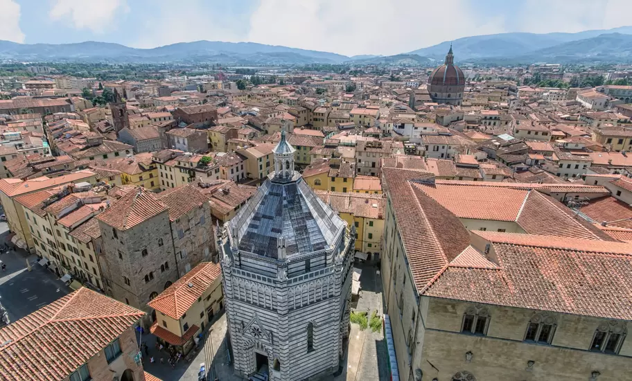 An aerial view over Pistoia, Italy, centred on an octagonal striped marble baptistery among red roofs, with hills beyond.