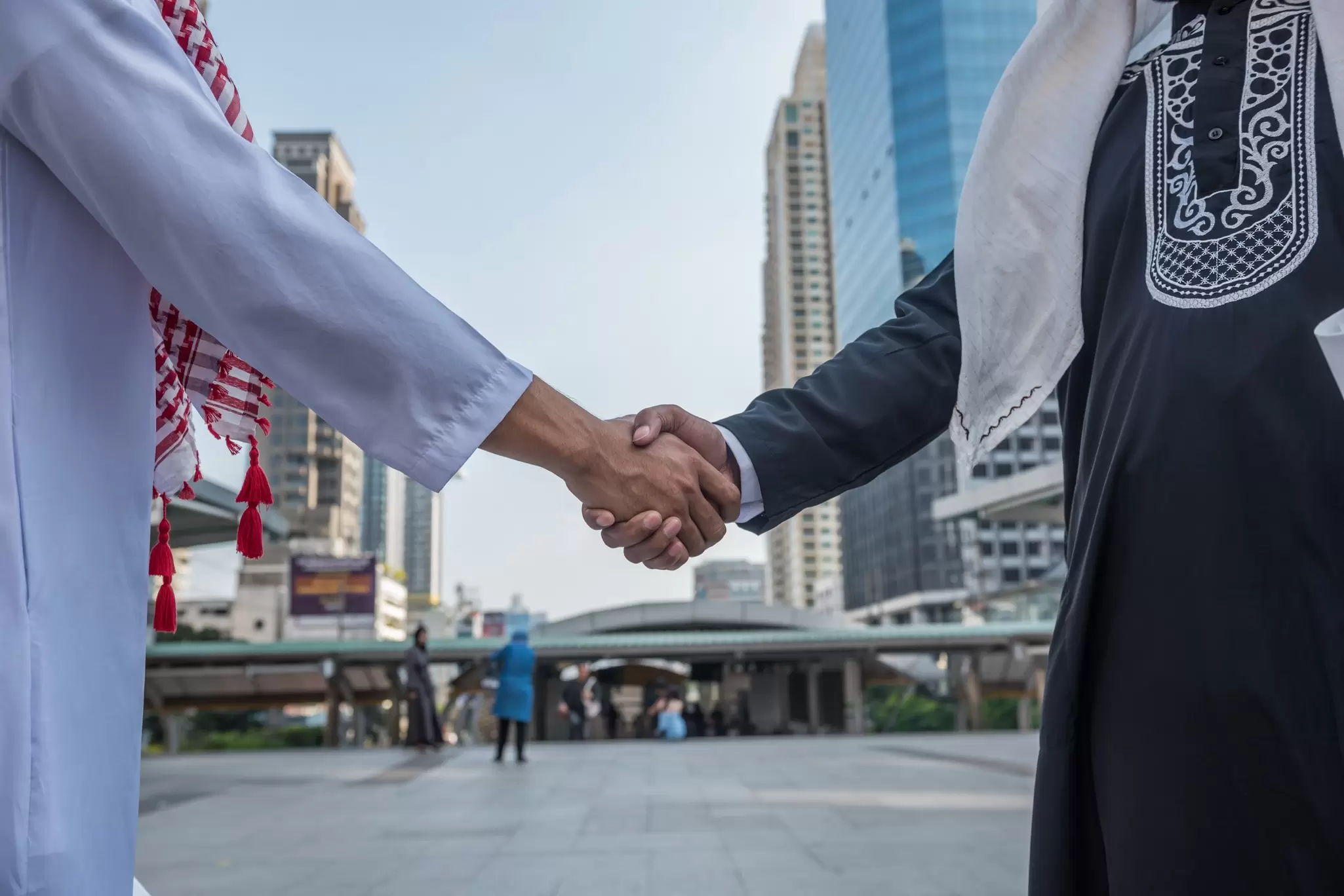 Two men in traditional Gulf Arab robes and headdresses shaking hands on a city plaza with skyscrapers behind
