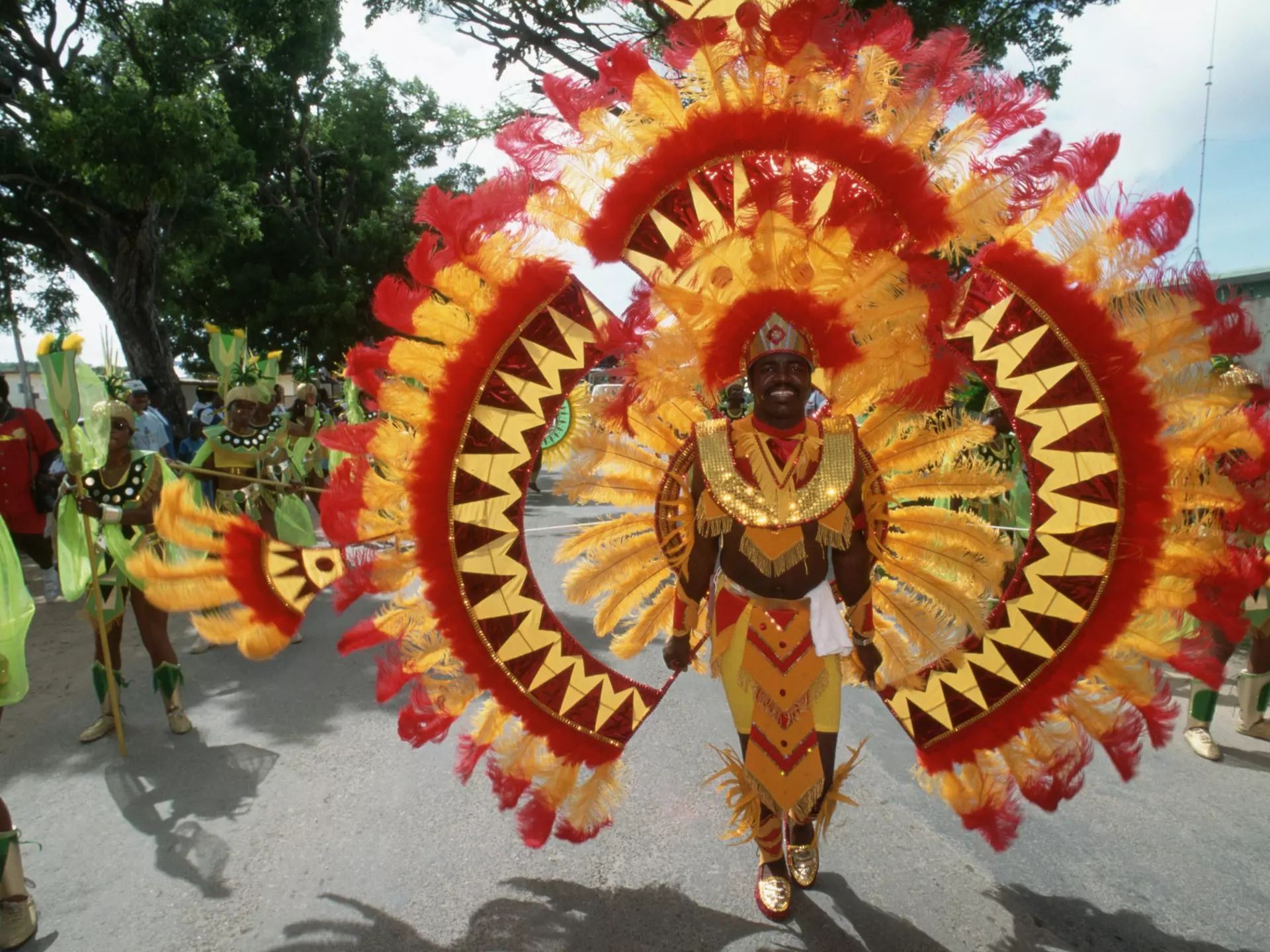A man participating in a street parade wears a red, yellow and orange outfit covered in feathers.