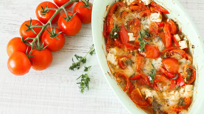 A vine with fresh red tomatoes and a casserole dish with squares of cheese topped with red peppers and green herbs.