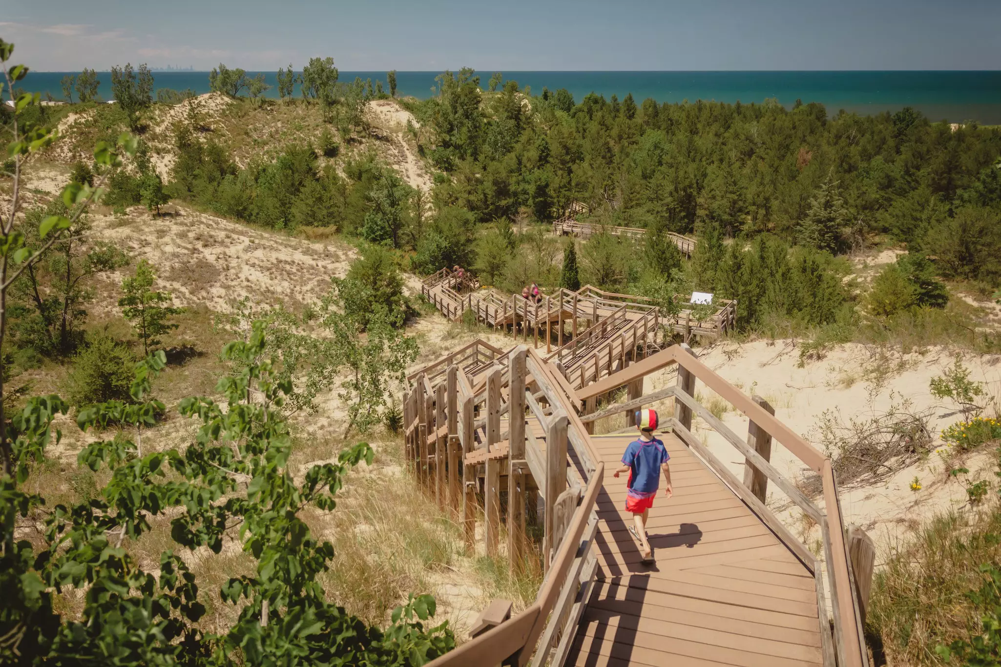 A child follows a boardwalk through sandy dunes and woodland towards the sea on a sunny day.