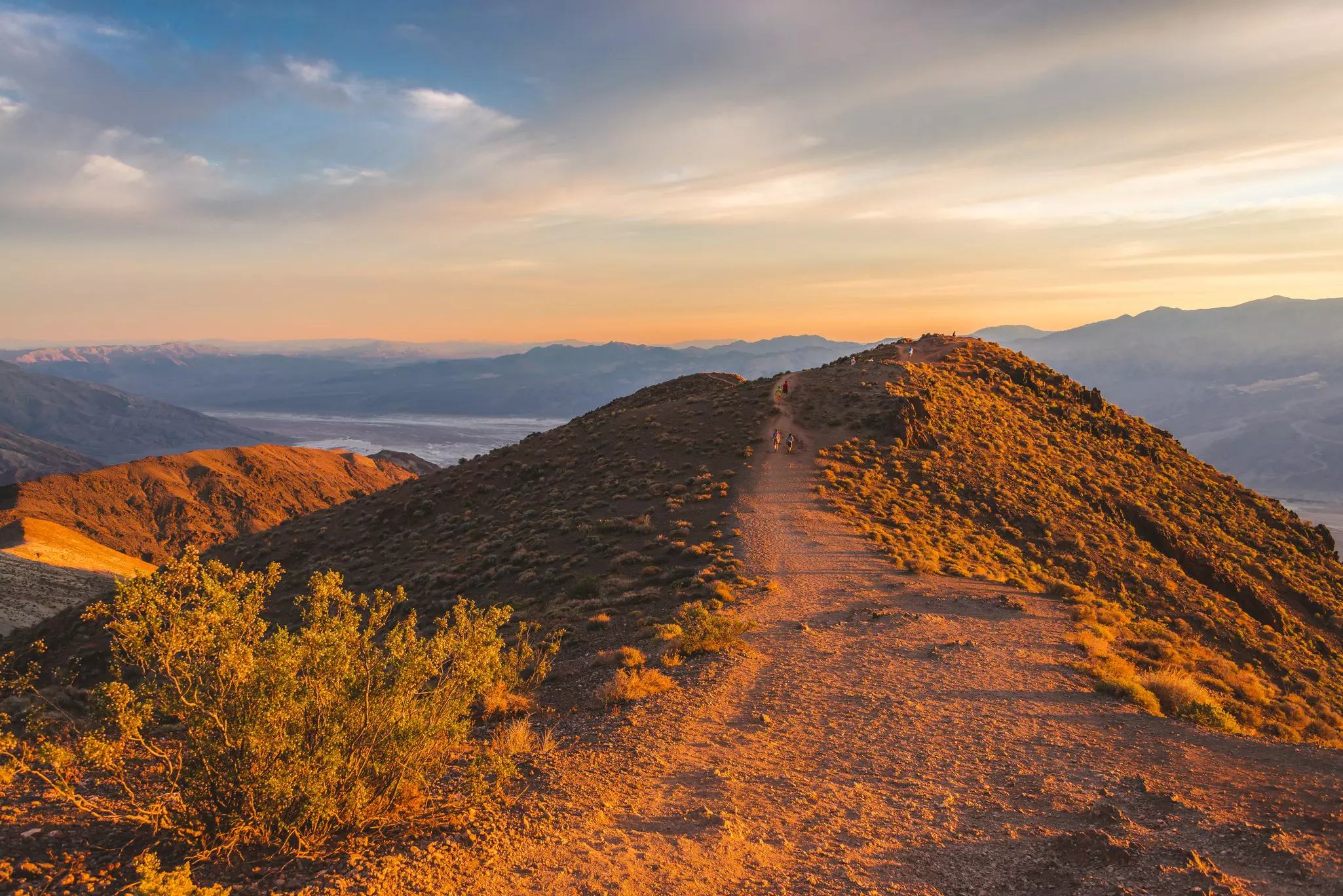 Hikers follow a dusty path up to a viewpoint from a peak