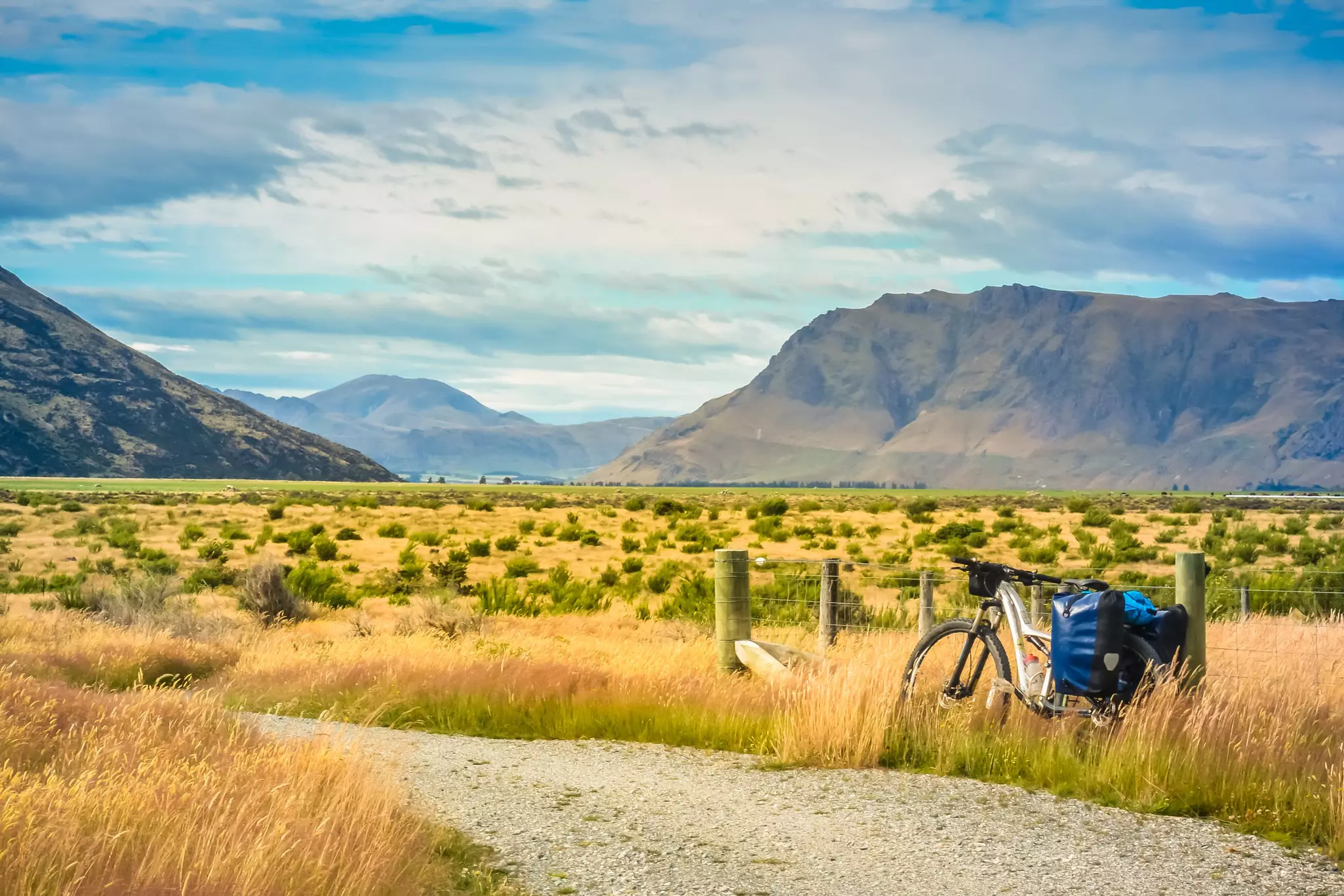 A bike with loaded panniers leans against a fence near a field in a mountainous area.