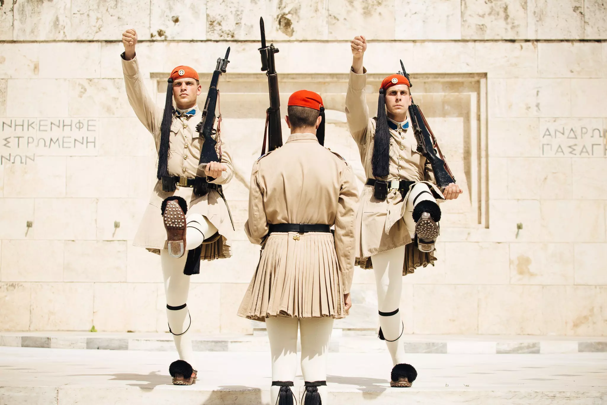 A guard wearing an earth-toned tunic and red beret and holding a bayonet in his left hand has his back toward the camera. Two identically dressed guards face him. Their right arms and left legs are raised, and they also carry bayonets in their left hands.