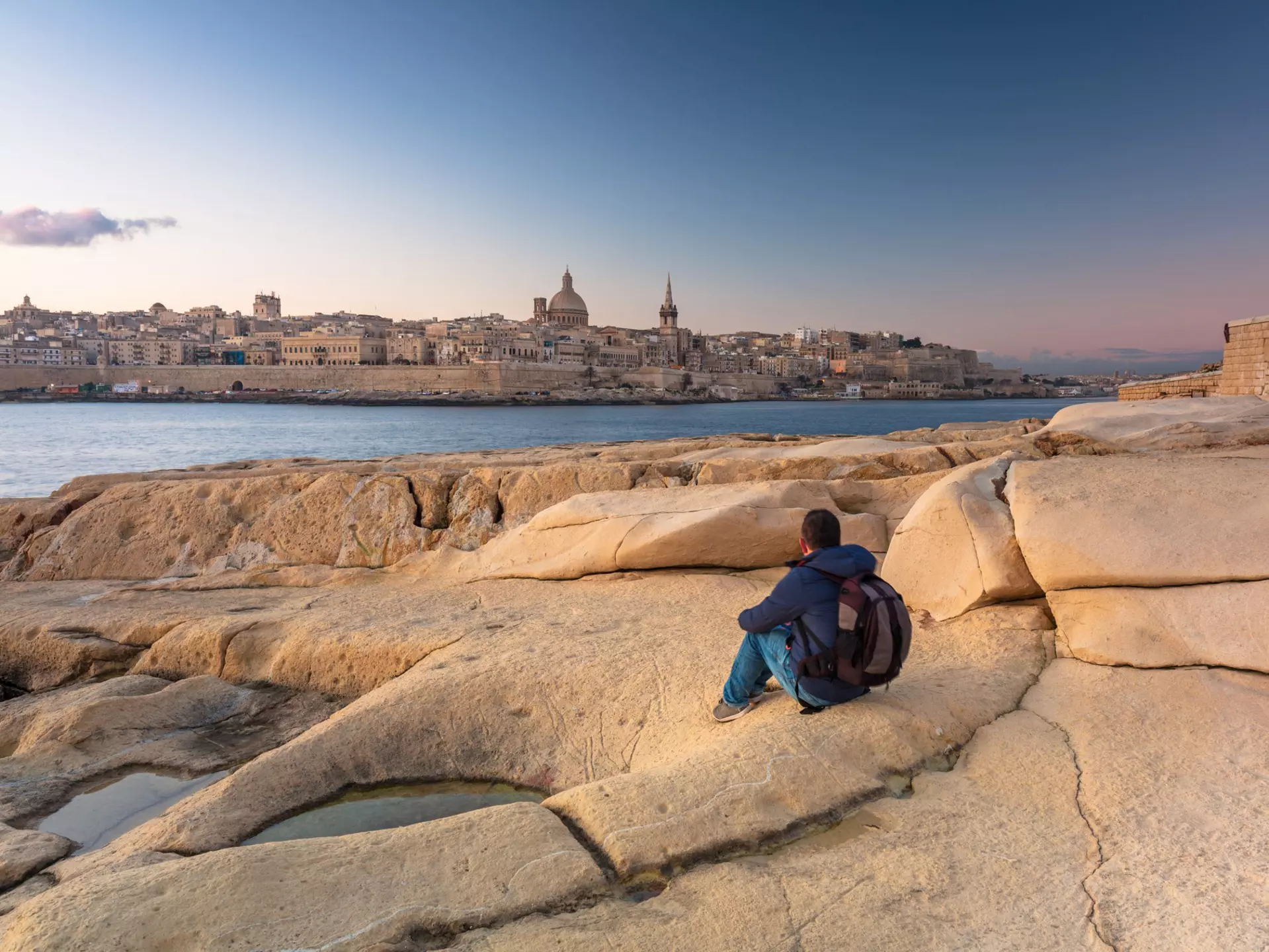 Man sitting on the rock and watch beautiful architecture of the Valletta city at dawn.