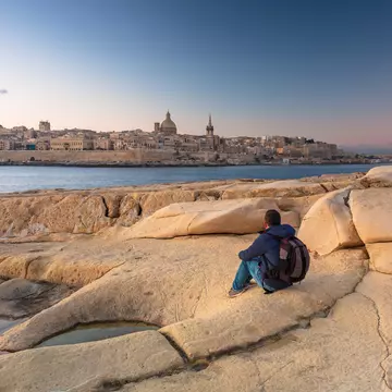Man sitting on the rock and watch beautiful architecture of the Valletta city at dawn.